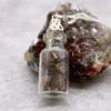 Tiger’s Eye Gemstone Bottle Pendant - Brown Crystal Charm Healing Crystals Gift sets. Small glass bottle pendant filled with polished brown tiger’s eye gemstones beside a rough crystal stone.