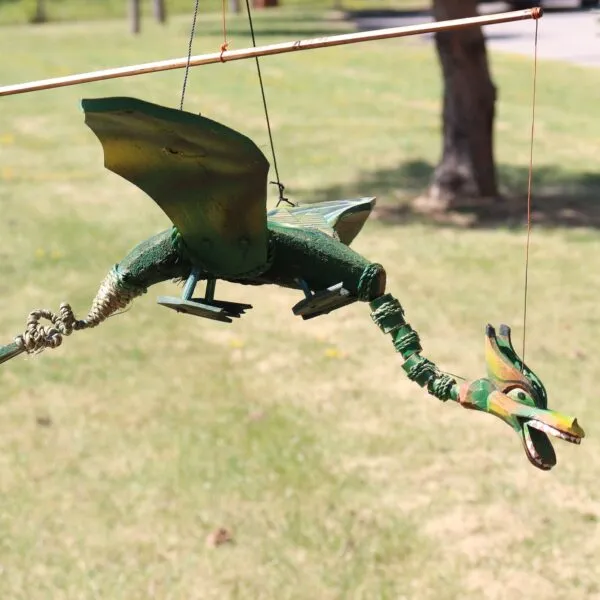 A colourful wooden dragon marionette hanging outdoors against a bright grassy park background.