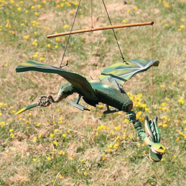 A handcrafted green dragon marionette with wings suspended outdoors above dry grass and yellow flowers.
