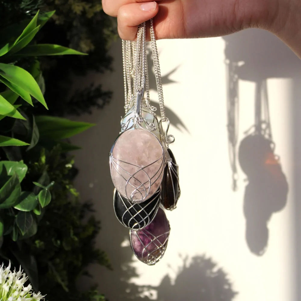 A hand holding silver-chain gemstone pendants with rose quartz, amethyst, and black stones in sunlight.