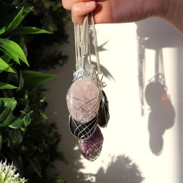 Hand holding silver wire-wrapped gemstone pendants, including a pink rose quartz, in natural sunlight.