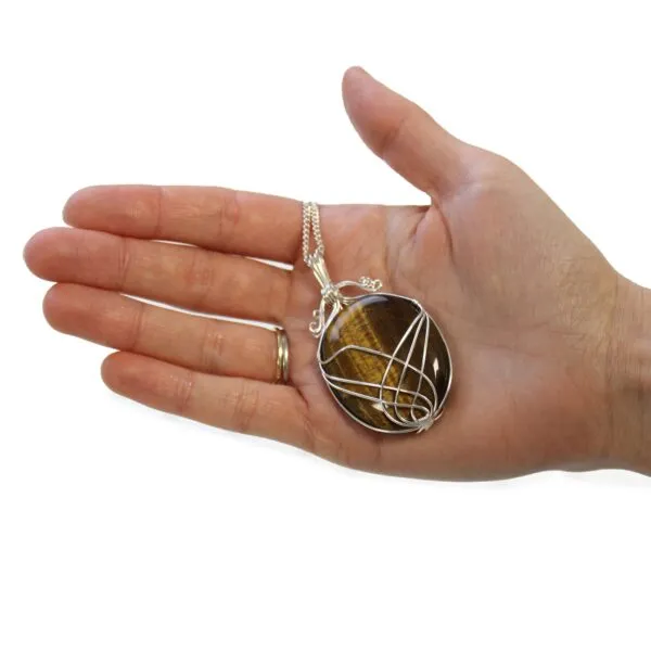 A silver wire-wrapped tiger’s eye gemstone pendant resting on an open adult hand.