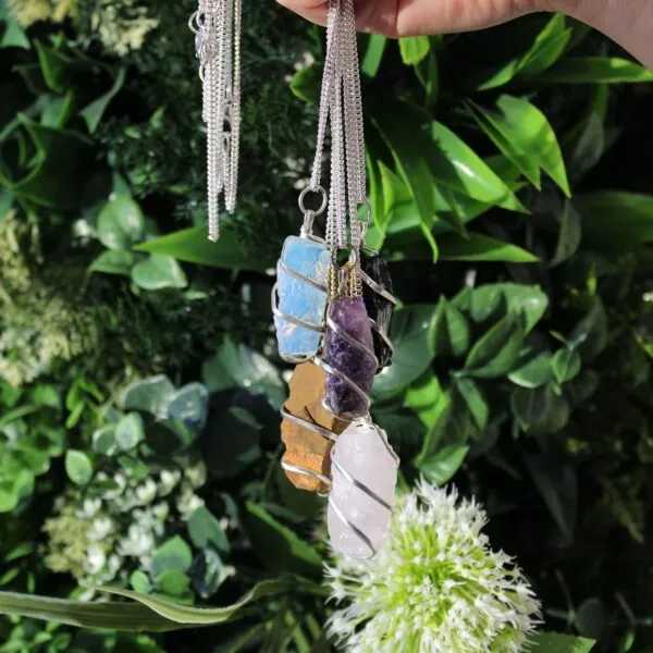 Hand holding silver wire-wrapped crystal pendant necklaces gleaming against a lush green garden background.