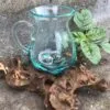 A hand-blown aqua glass pitcher resting on sculpted driftwood beside a small green plant.