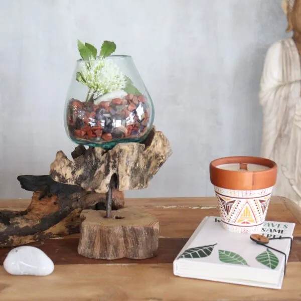 A wooden table with a glass plant arrangement, terracotta candle pot, white stone, and Buddha statue.