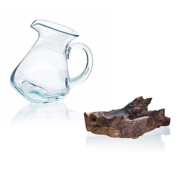 A clear handmade glass pitcher beside textured driftwood on a white background.