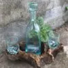 Artisanal Glass Bottle And Glasses Set - Rustic Driftwood Base Clear artisanal glass bottle and glasses displayed on rustic driftwood with green leafy plant backdrop.