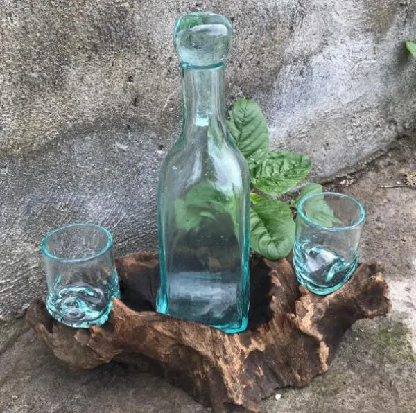 Artisanal Glass Bottle And Glasses Set - Rustic Driftwood Base Clear artisanal glass bottle and glasses displayed on rustic driftwood with green leafy plant backdrop.