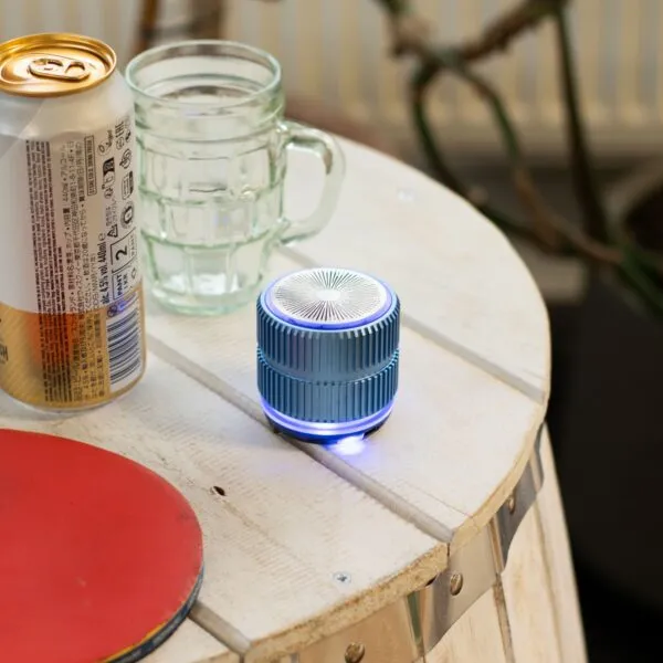 A blue portable Bluetooth speaker on a wooden table with a drink can, glass, and ping pong paddle.