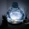 A glowing translucent Laughing Buddha statue with a succulent and reed diffuser on a woven surface.