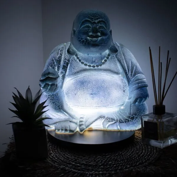 A glowing translucent Laughing Buddha statue with a succulent and reed diffuser on a woven surface.