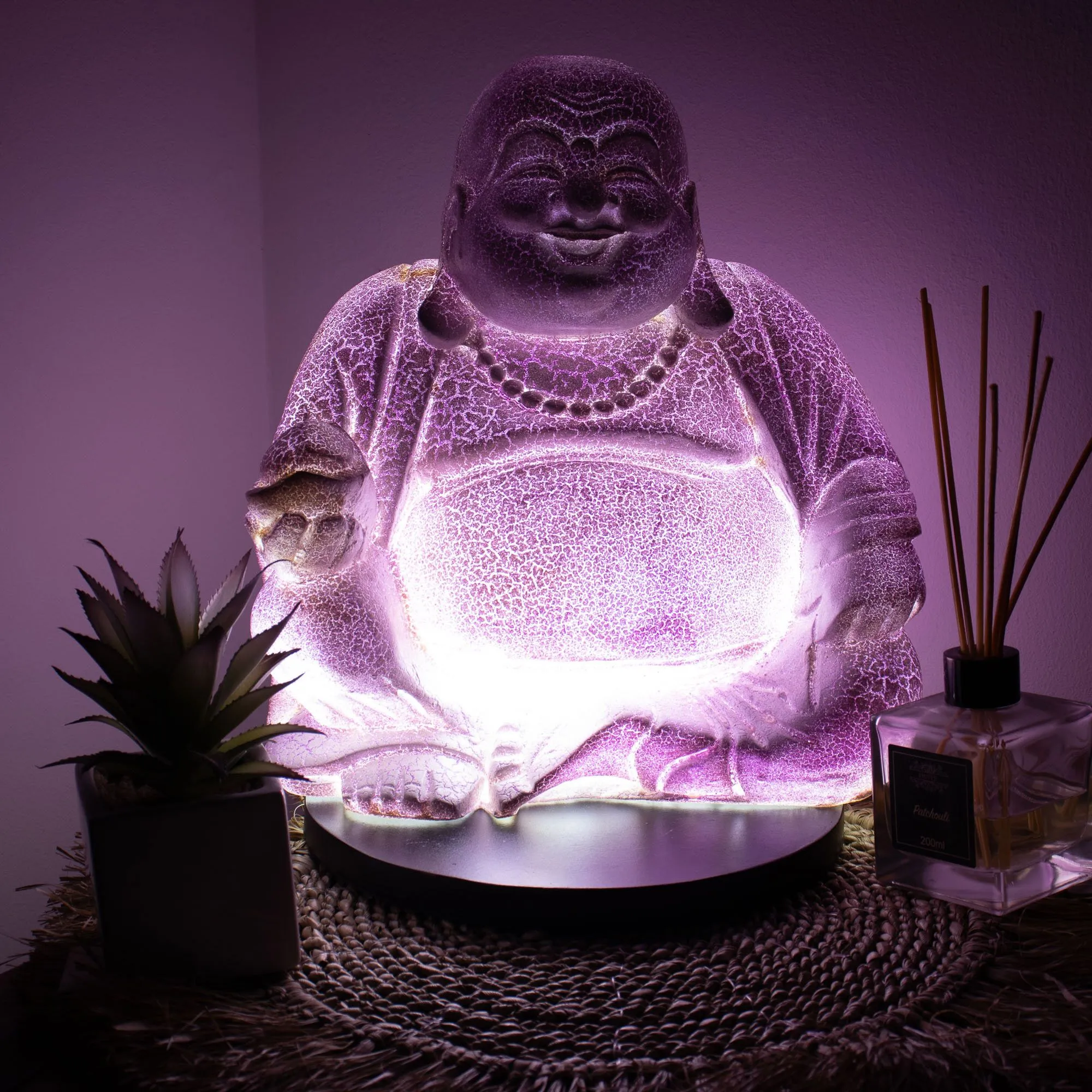 A glowing purple Laughing Buddha lamp beside a succulent plant and patchouli reed diffuser.