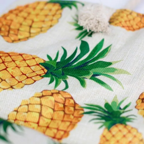 Textured beige burlap fabric featuring a tropical pineapple print and decorative cotton tassel.