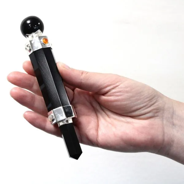 Hand holding a black obsidian obelisk wand with silver accents and orange stones.